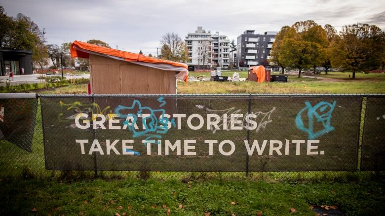 A chain-link fence surrounds an empty lot with a banner hanging on the fence that says great stories take time to write.