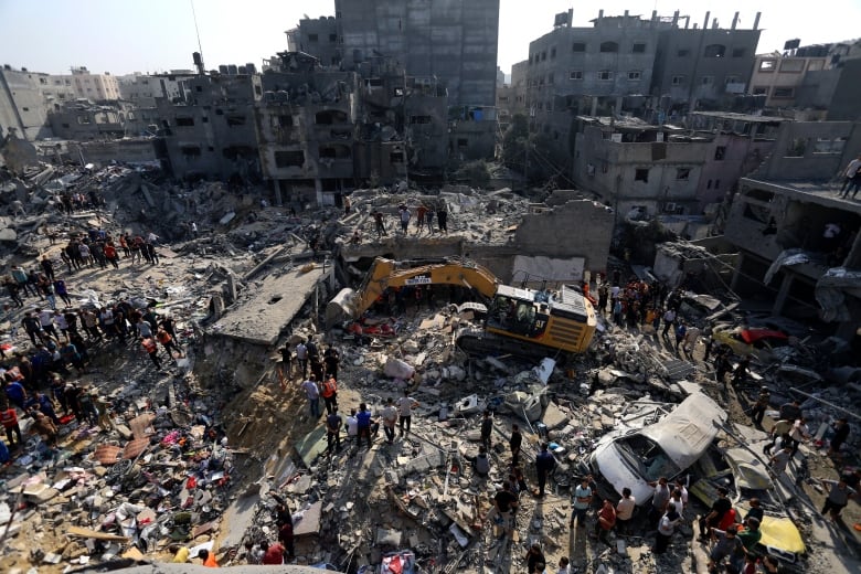 A wide view of many destroyed concrete buildings surrounding a people and equipment sifting though rubble. 