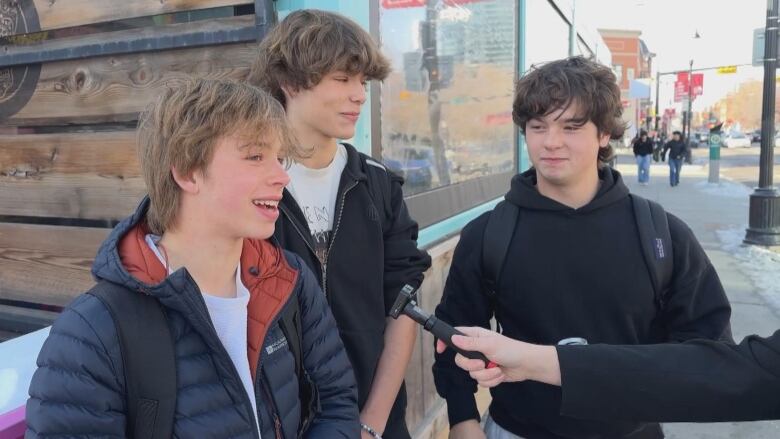 three young boys stand outside, one speaks into a microphone