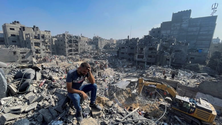 Holding one hand to his head, a distraught man sits on rubble overlooking an area hallowed out by airstrikes.