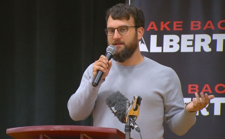 A man wearing a grey long-sleeved shirt speaks into a microphone in front of a podium.