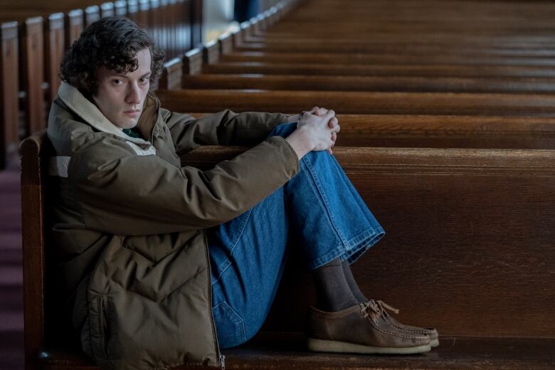 A young man is seated in a church pew. His feet are up on the bench as he leans against the arm rest. 