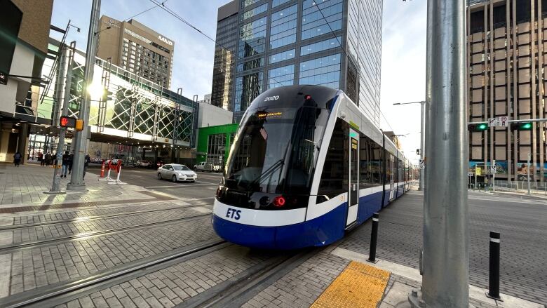 A blue and white train rolls across some tracks. Various buildings are in the background.