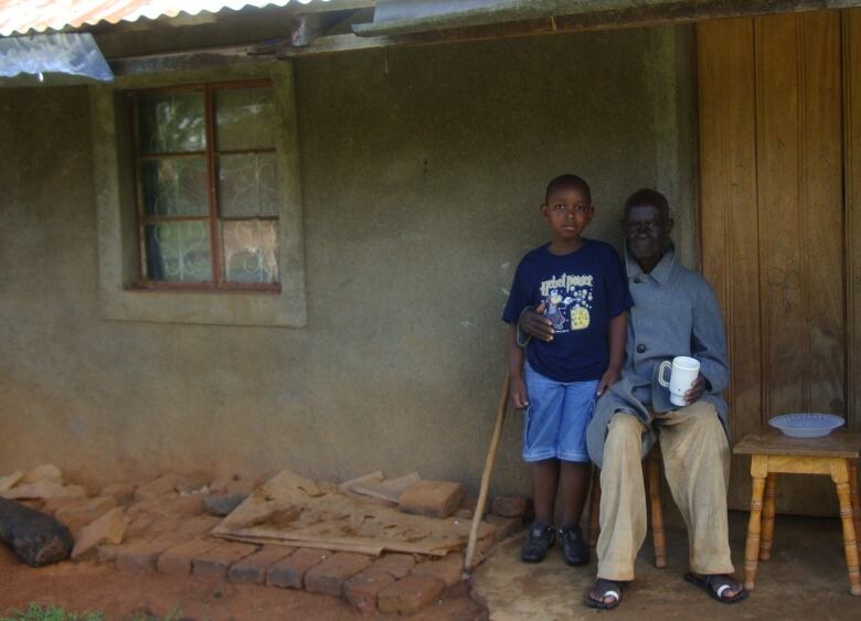 A boy wearing shorts and a T-shirt stands beside an older man sitting on a stool and holding a coffee cup. The two are posed outside a building with rough walls and a tin roof.