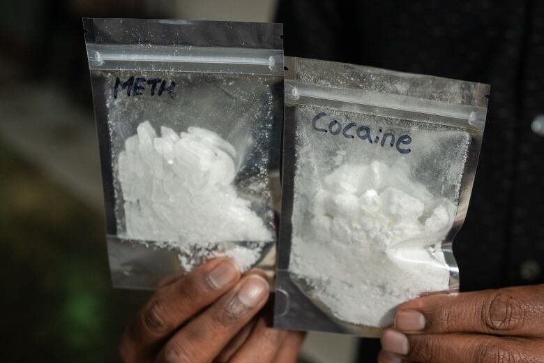 Two hands hold two bags of white powder drugs, one labelled meth, the other labelled cocaine.