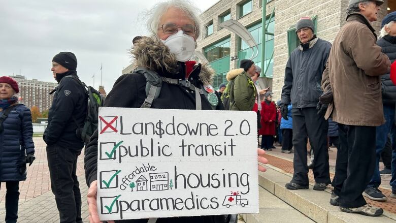 Woman holds a sign that says X to Lansdowne and checkmark to public transit, housing and paramedics