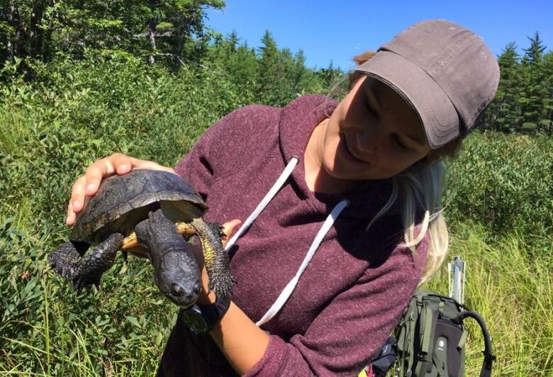 A woman standing in front of an area with green shrubs and bushes. She is holding a Blanding's turtle.