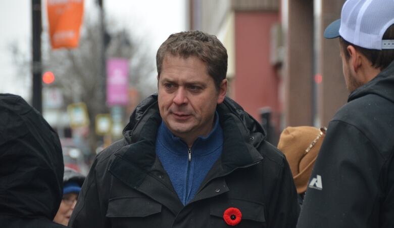 A man wearing a dark jacket with a poppy on it frowns while standing on the street