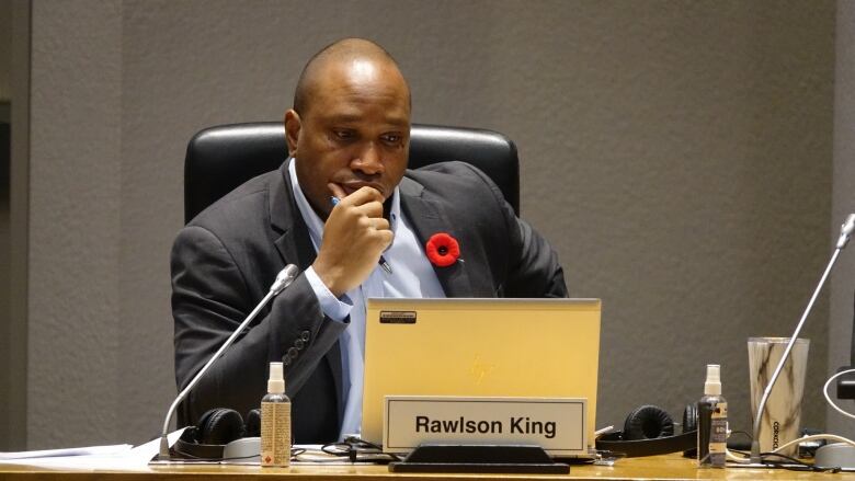 A man holding his chin looks down at his laptop. He's wearing a red poppy pin on a dark suit.