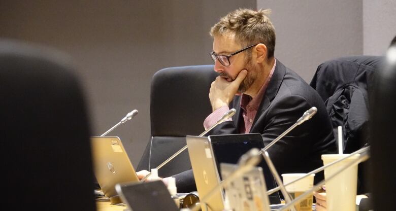 Side view of a man on his computer, touching his chin with one hand. A line of laptops seen below him.