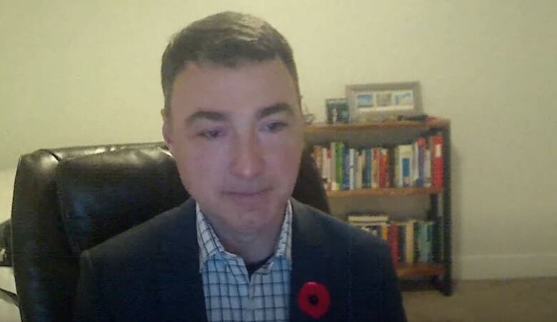 A man with short brown hair, wearing a suit jacket, sits in a room with a bookcase behind him.