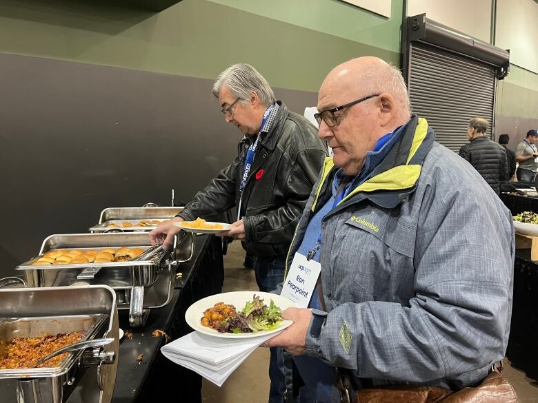 A man wearing a jacket with a lanyard holding a plate full of food stands at a buffet line.