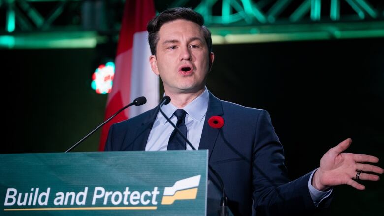 A man in a suit speaks at a podium, with a sign reading 