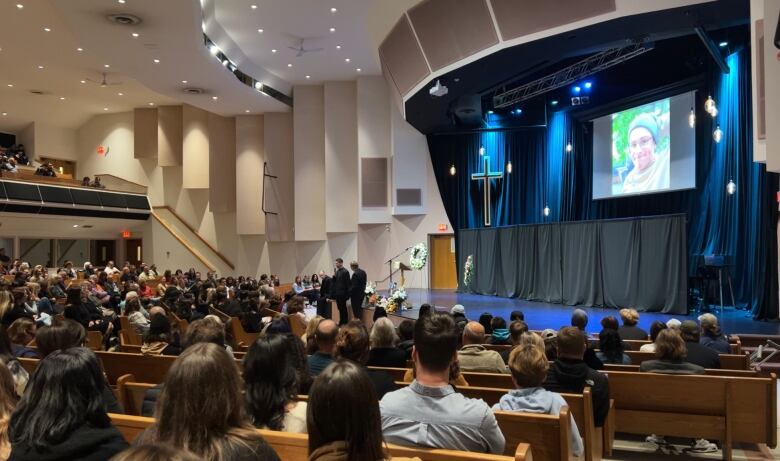 People sit in pews at a church as two men stand on stage speaking. A large photo of a young teen is on the screen. 