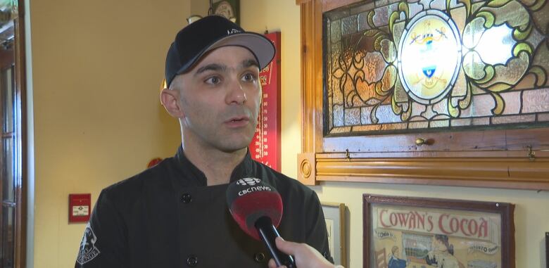 A man in a dark chef's coat and baseball cap stands in front of a stained-glass window, talking into a microphone. 