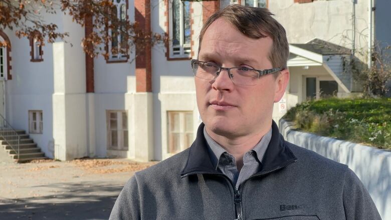 A white man with glasses and brown hair wearing a grey sweater stands outside with a large white building behind him