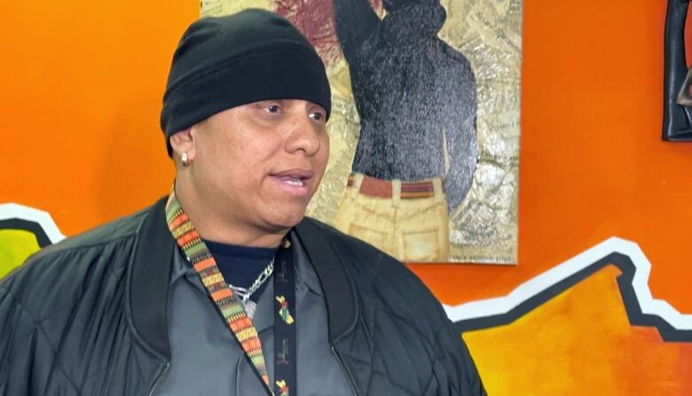 A Black man with a black fitted toque and black jacket stands in front of a bright orange wall with artwork hanging behind him 