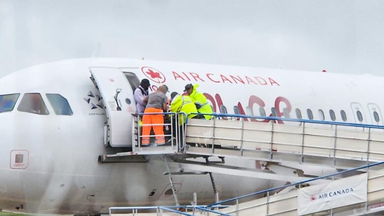 A long distance shot of multiple workers crowded around Alessia in her wheelchair outdoors on a ramp. 