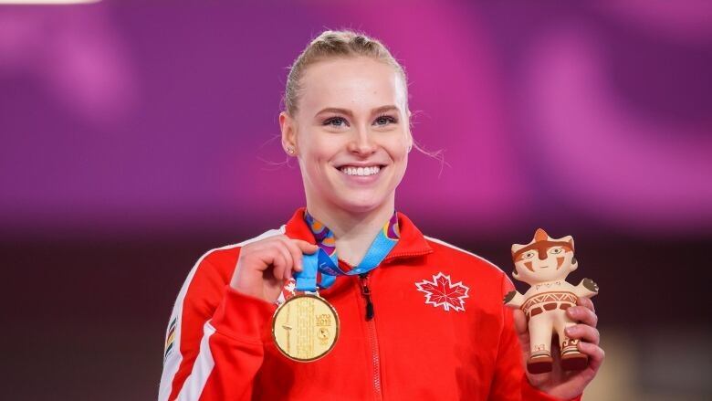 A young woman wearing a read tracksuit sweater smiles and holds up a gold medal that hangs around her neck.