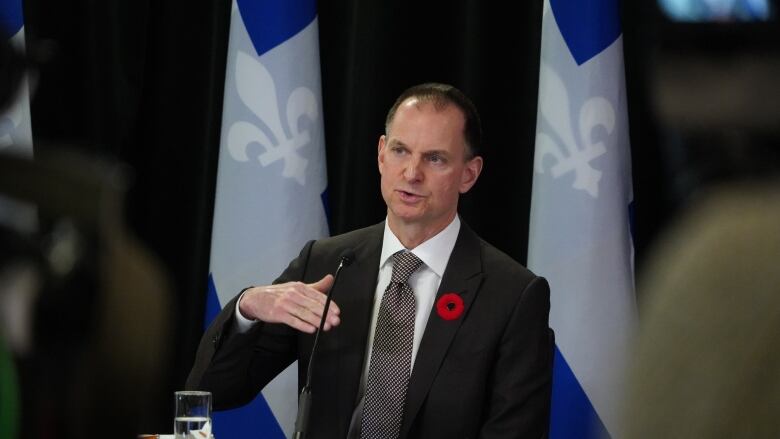 Man in suit gestures in front of microphone. Blue and white flags hang behind him.