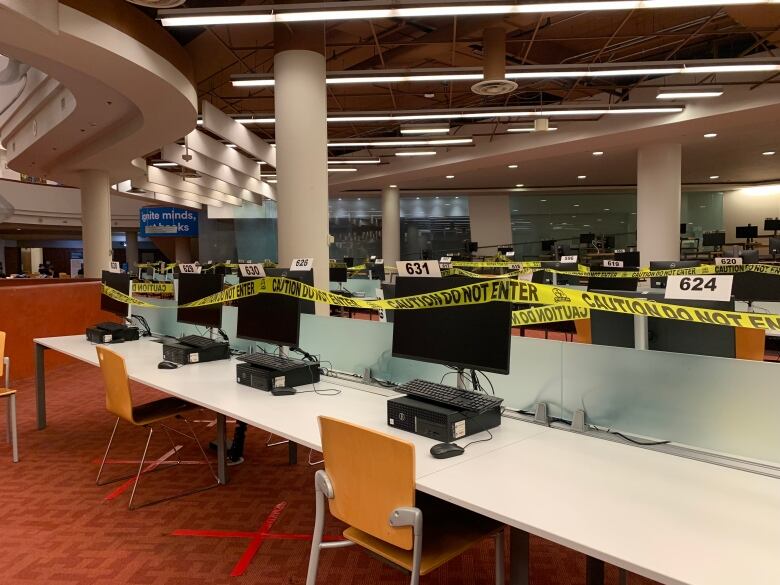 A row of desktop computers in an empty library are roped off by yellow tape.