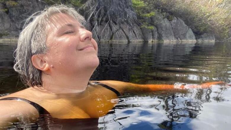 A woman in a body of water with her arms floating on each side of her. She has grey hair and her face is tilted up with her eyes closed