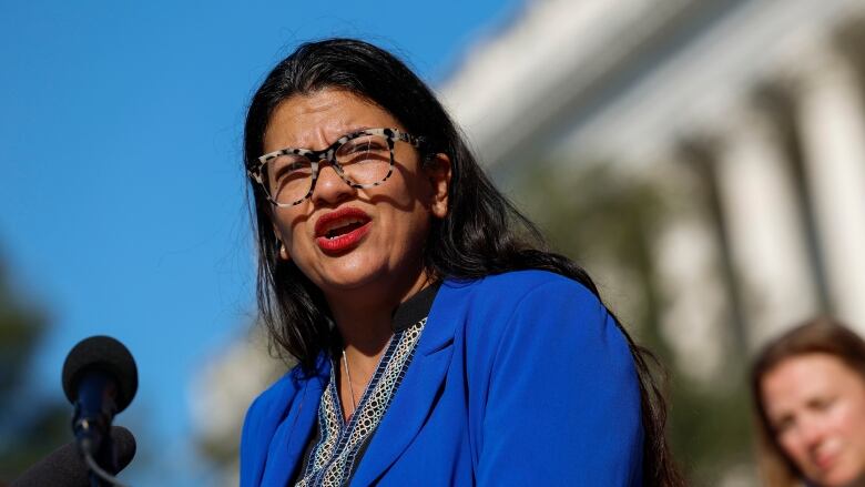 A closeup of a bespectacled woman wearing a blazer speaking while outdoors is shown.