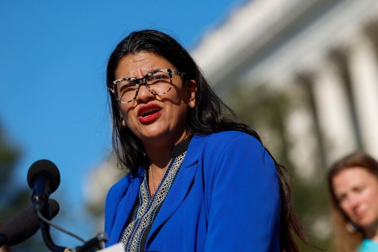 A closeup of a bespectacled woman wearing a blazer speaking while outdoors is shown.