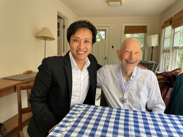 A man with black short hair wearing a black blazer and a bald man wearing a medal around his neck put their arms around each other and smile at a table.