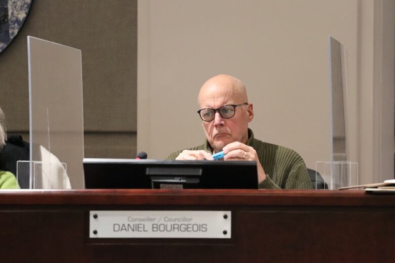 A frowning bald man in a green sweater sitting behind a large desk with a computer screen holding a blue highlighter. 