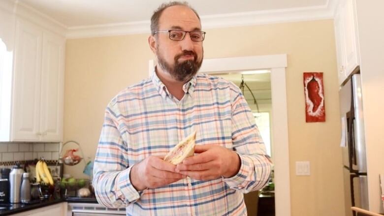 A man with glasses and a button-up shirt looks at the camera before taking a bite of a sandwich.