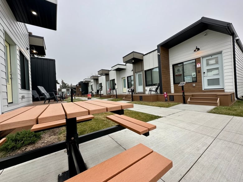 brown picnic table in the foreground, next to a sidewalk and white and brown tiny homes lining the walk. 