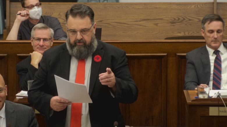 A man with a dark suit and red tie standing up pointing at politicians on the other side of the aisle.