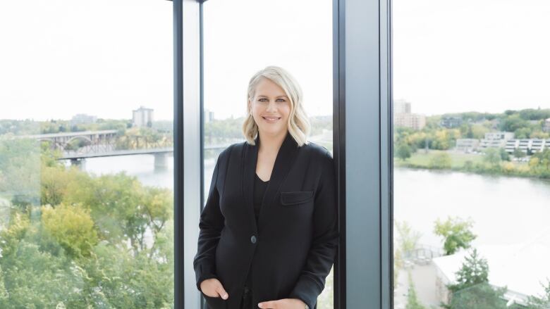 A blonde woman in an all black outfit sits on a chair. A high top view behind her shows sky and a tall building