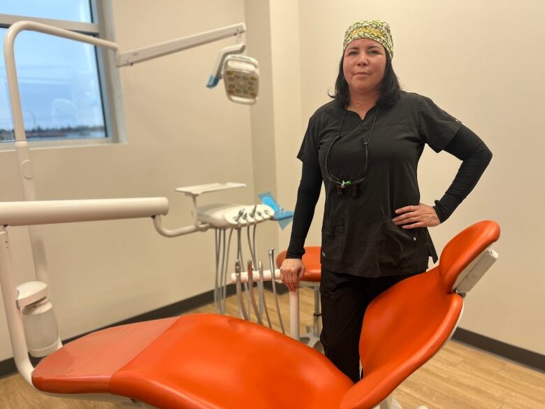 A woman can be seen standing in a dentists office, she is wearing black scrubs. 