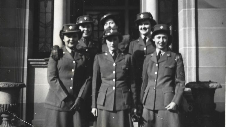 Six smiling young women in military clothing stand on some stone steps in an image from the 1940s. 