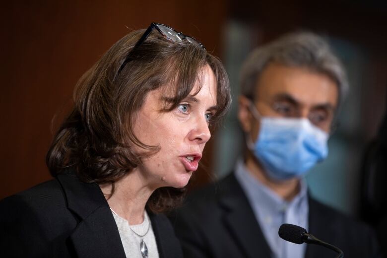 A white woman with short brown hair speaks next to a South Asian man wearing a mask.