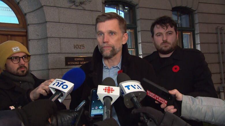 A man in a coat speaks to reporters holding microphones.  