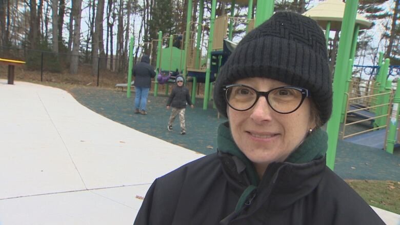 A woman wearing a toque and glasses stands near a playground. People can been seen playing in the background.