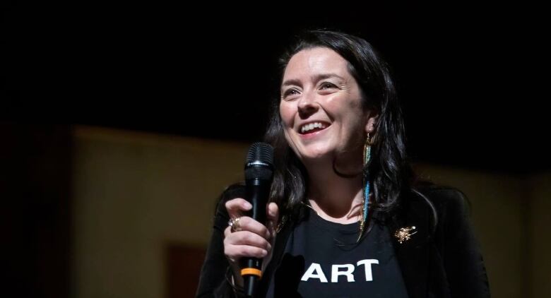 A smiling woman holds a microphone. She is wearing a black T-shirt with white letters that say 