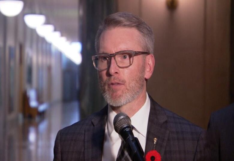 A man wearing glasses, with a grey beard, and wearing a suit, speaks in front of a microphone.