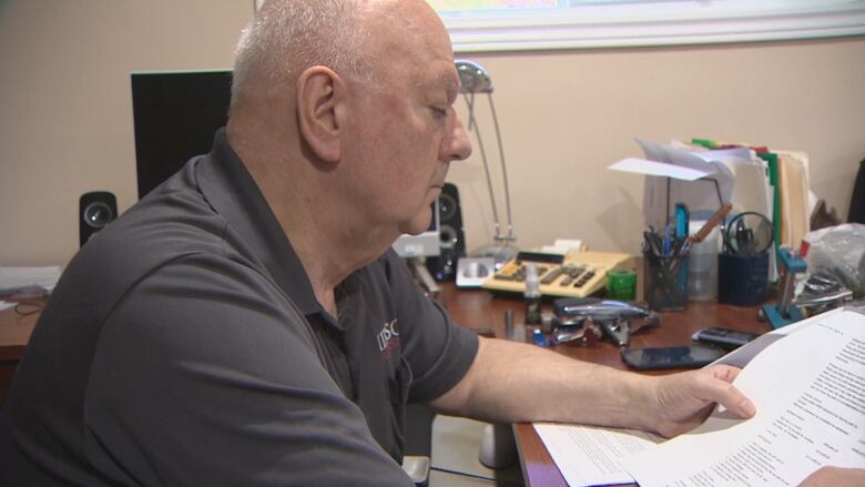 A bald man in a dark polo shirt sits at a desk and looks over documents. 