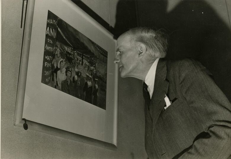 Famous painter Arthur Lismer is shown in a black and white photo looking at a piece of artwork.