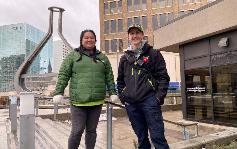 A woman wearing braids and a green jacket and a man with a baseball hat and black jacket are pictured in downtown Winnipeg.