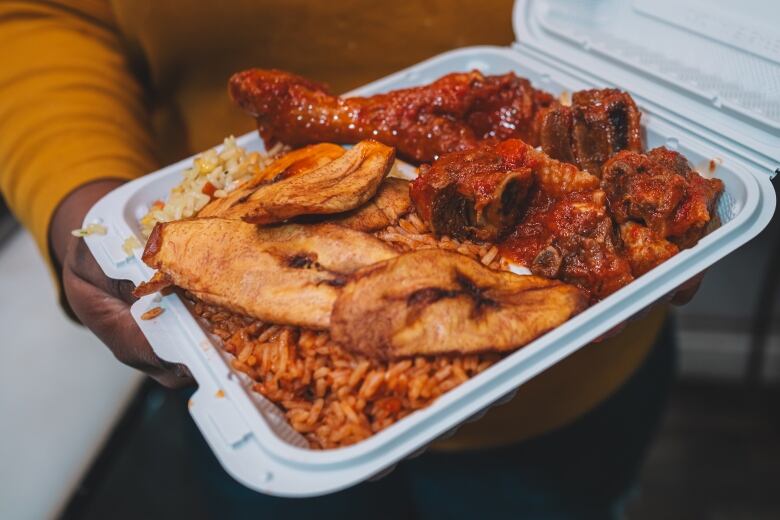 A takeout container filled with two types of rice, fried plantain, and meat in a deep red sauce.