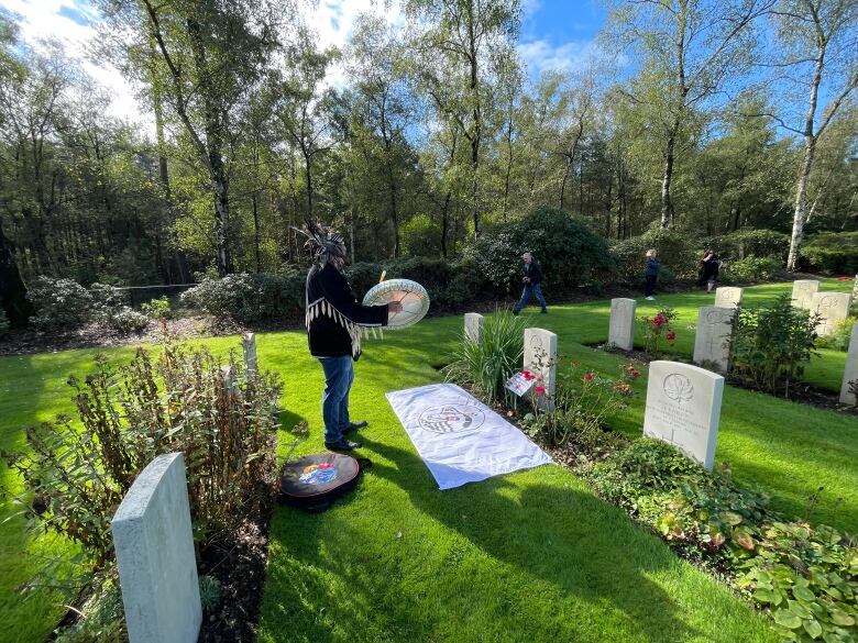 A man stands at a grave, beating a drum. 