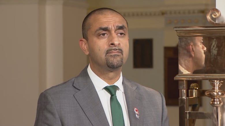 A man with a close shaved head and goatee looks to the right of the frame. He's wearing a grey suit, white shirt, and green tie.