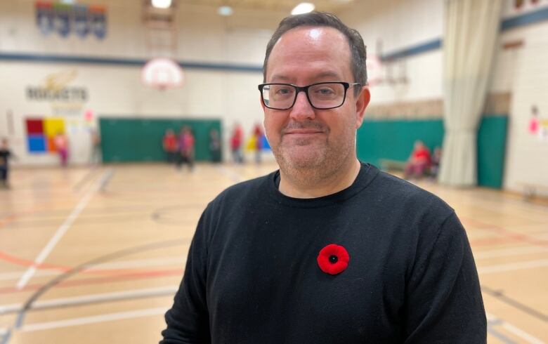 A man in a black sweater with a poppy pinned on it stands in a school gymnasium.