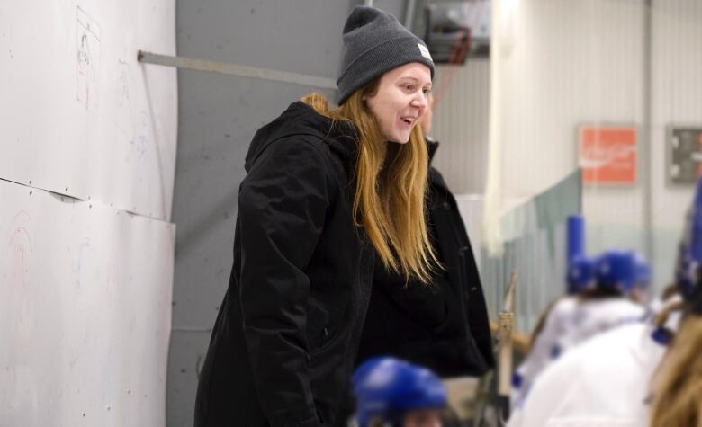 A smiling woman in a tuque stands behind hockey players sitting on a bench.