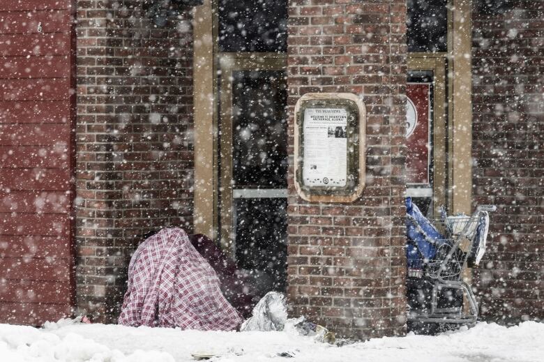 A homeless person sits under a blanket outside a building during a heavy snowstorm.